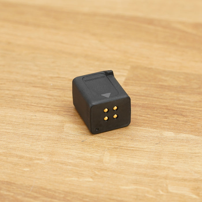 A small black rectangular Shimano Di2 battery photographed on a light brown wooden surface against a plain white wall.
