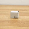 A small square off-white cardboard box with black printed text and a QR code, sitting on a light brown wooden surface.
