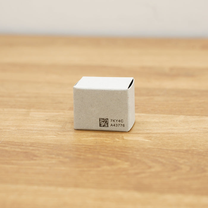 A small square off-white cardboard box with black printed text and a QR code, sitting on a light brown wooden surface.