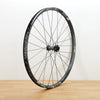A black aluminum DT Swiss front wheel photographed on a light brown wooden surface against a plain white wall.