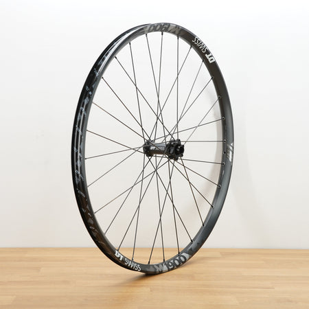 A black aluminum DT Swiss front wheel photographed on a light brown wooden surface against a plain white wall.
