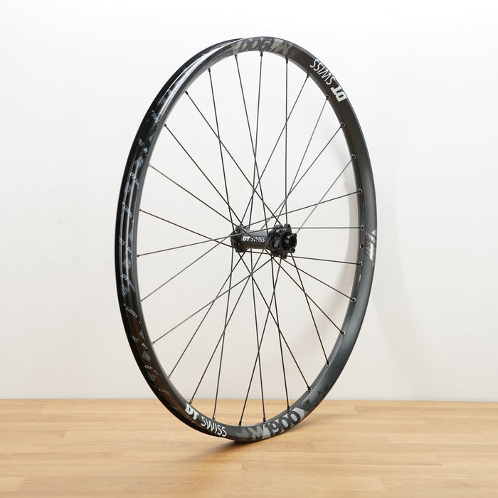 A black aluminum DT Swiss front wheel photographed on a light brown wooden surface against a plain white wall.