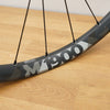 Close-up of a bicycle wheel rim with black spokes, featuring a dark gray and white geometric pattern and the text 'M1900' on the rim, resting on a light wood floor.