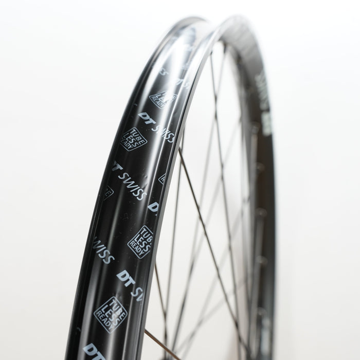 Close-up of a black DT Swiss bicycle wheel rim with white printed branding and thin silver spokes against a plain white background.