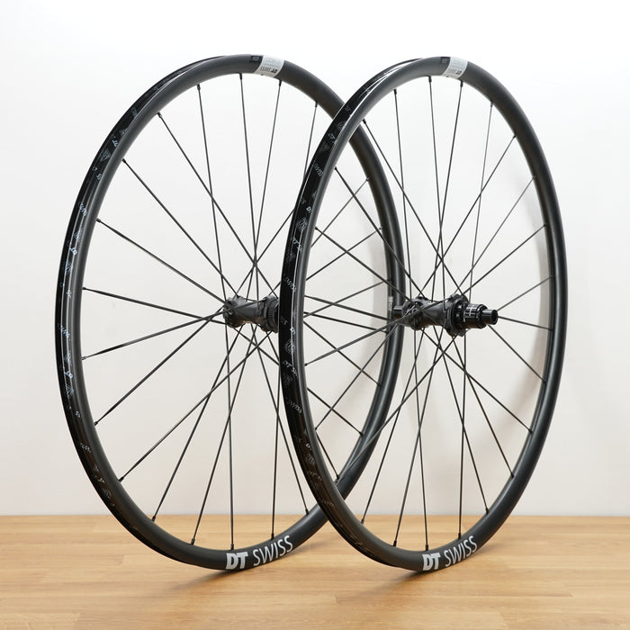Two black DT Swiss aluminum road bicycle wheels standing upright on a light wood surface against a plain white wall.