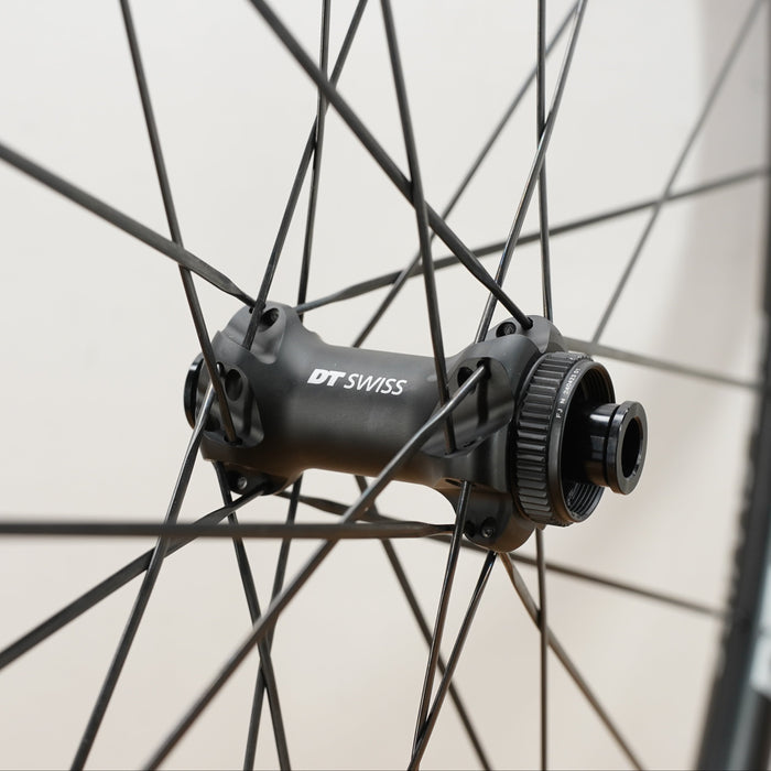 Close-up of a black DT Swiss wheel hub mounted on a bicycle wheel, with thin black spokes and part of the tire visible against a light neutral background.