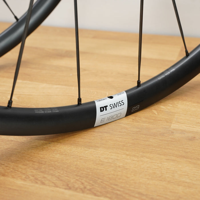 Close-up of a black DT Swiss E1800 road bike wheel with thin black spokes, resting on a light wood surface.