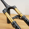 A close-up of a mountain bike front suspension fork with black and gold components, resting on a wooden floor against a plain wall.
