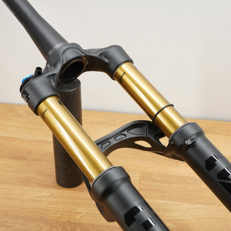 A close-up of a mountain bike front suspension fork with black and gold components, resting on a wooden floor against a plain wall.
