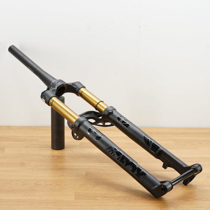 A black and gold mountain bike front suspension fork displayed on a wooden table against a plain white wall.