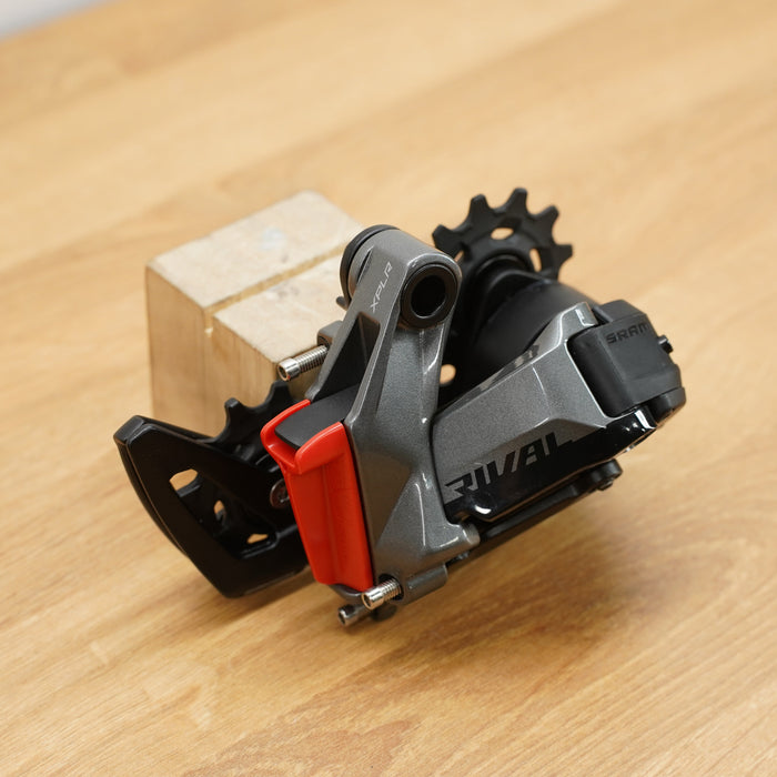A grey and black SRAM bicycle rear derailleur with a red battery cover photographed on a light brown wooden surface against a plain white background.