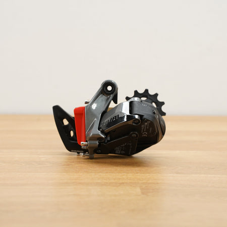 A grey and black SRAM bicycle rear derailleur with a red battery cover photographed on a light brown wooden surface against a plain white background.