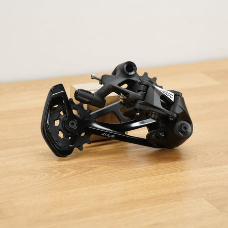 A black SRAM rear derailleur component with visible SRAM branding, resting on a light wood surface against a plain off-white background.