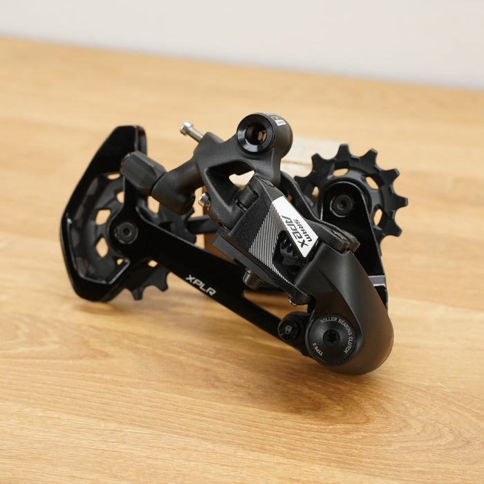 A black SRAM rear derailleur component with visible SRAM branding, resting on a light wood surface against a plain off-white background.