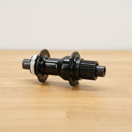 A Shimano black bicycle hub with silver metal splines, resting on a light wood tabletop against a neutral off-white background.