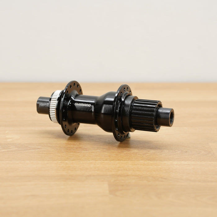 A Shimano black bicycle hub with silver metal splines, resting on a light wood tabletop against a neutral off-white background.