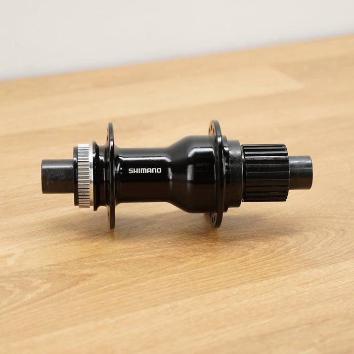 A Shimano black bicycle hub with silver metal splines, resting on a light wood tabletop against a neutral off-white background.