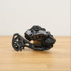 A black and dark gray Shimano GRX bicycle rear derailleur resting on a light wood tabletop against a plain off-white wall.