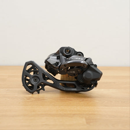 A black and dark gray Shimano GRX bicycle rear derailleur resting on a light wood tabletop against a plain off-white wall.