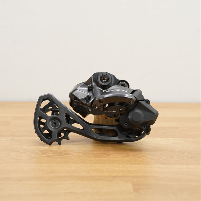 A black and dark gray Shimano GRX bicycle rear derailleur resting on a light wood tabletop against a plain off-white wall.