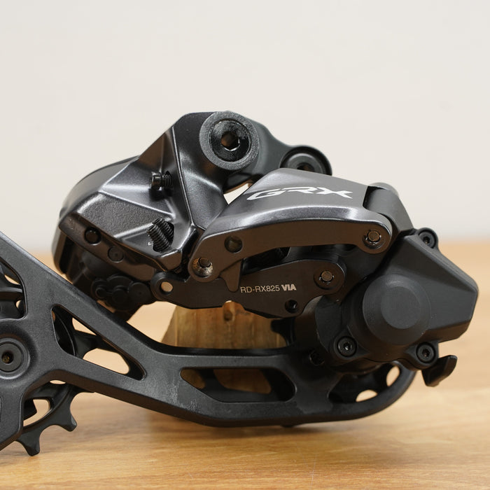 A close-up photo of a black and dark gray Shimano GRX bicycle rear derailleur resting on a light wood tabletop against a plain off-white wall.