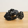 A black bicycle rear derailleur on a wooden surface against a plain white wall.
