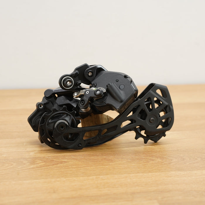 A black bicycle rear derailleur on a wooden surface against a plain white wall.