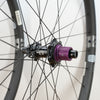 Close-up of a bicycle wheel hub with a prominent purple anodized freehub, black spokes, and a dark gray rim against a plain white background.