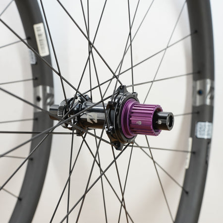 Close-up of a bicycle wheel hub with a prominent purple anodized freehub, black spokes, and a dark gray rim against a plain white background.