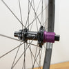 Close-up of a bicycle hub with a black body and a metallic purple splined end, mounted on a wheel with thin black spokes against a light wall.